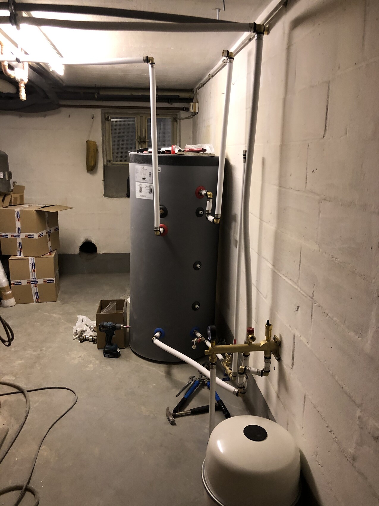 Großer runder Warmwasser-Boiler im Keller, umgeben von Rohren, Werkzeugen und Kartons.