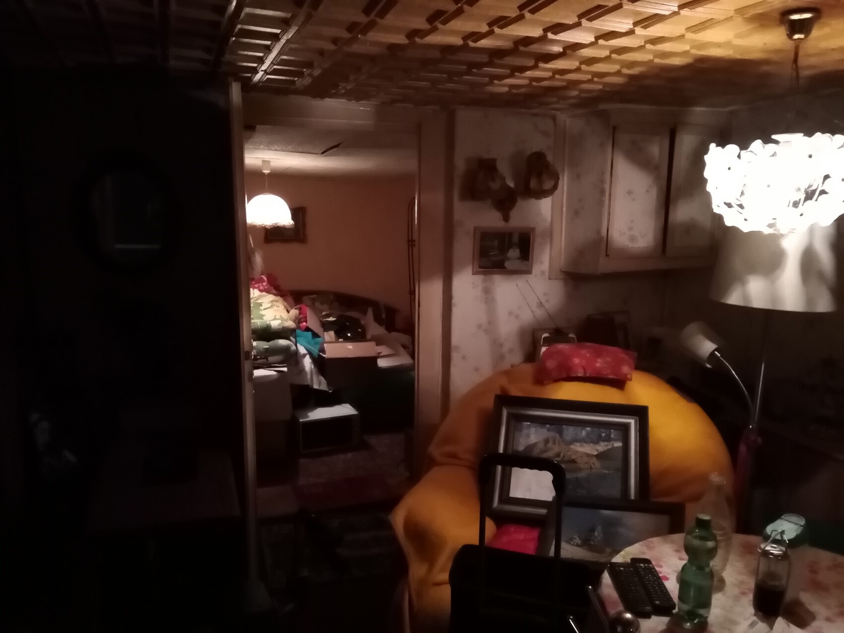 Dunkles Kellerzimmer mit Moebelchaos, Lampe und Esstisch im Vordergrund.