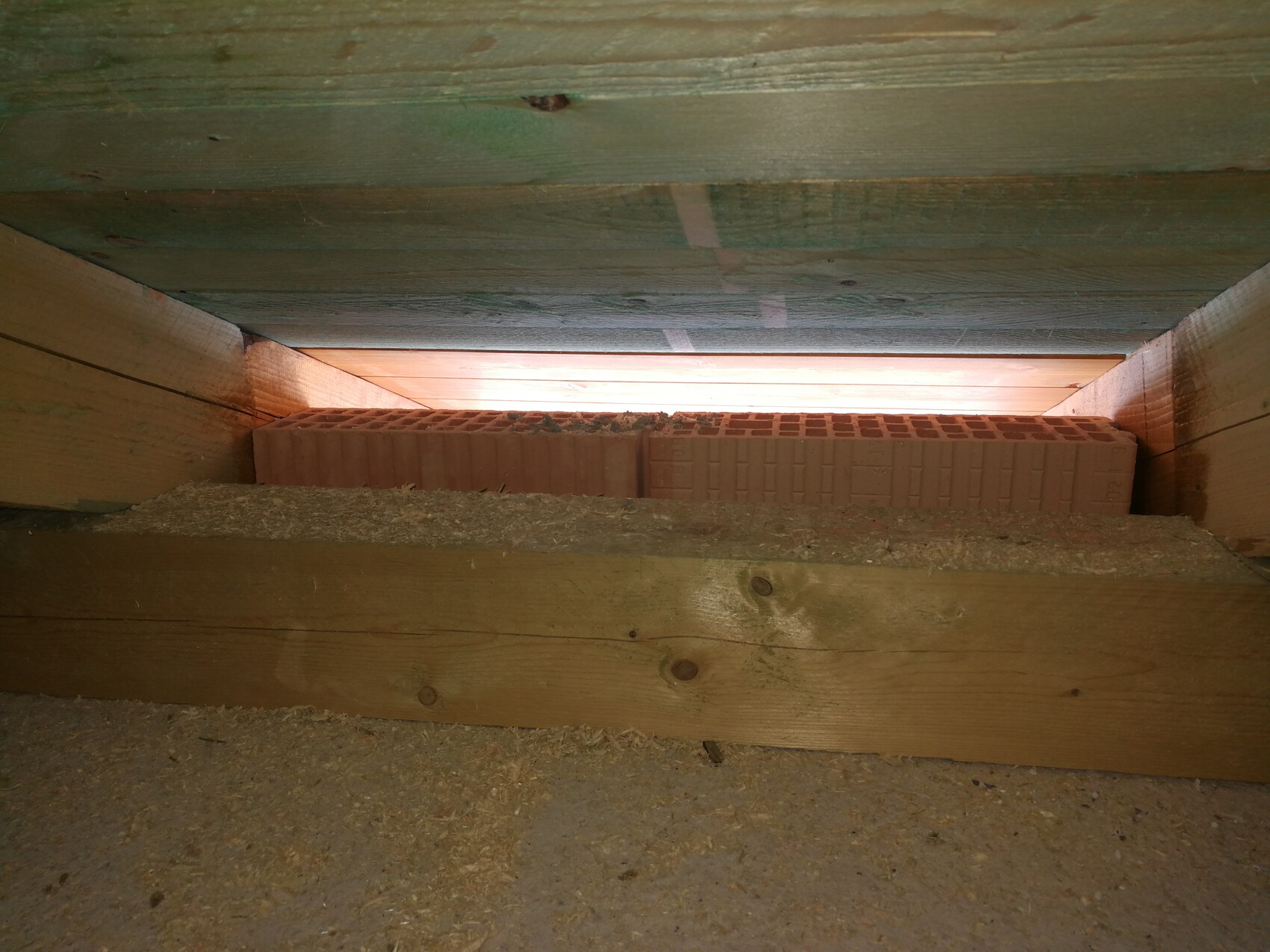 Roter Ziegel liegt quer zwischen Holzbrettern in einer engen Dachbodenöffnung, Staub am Boden.