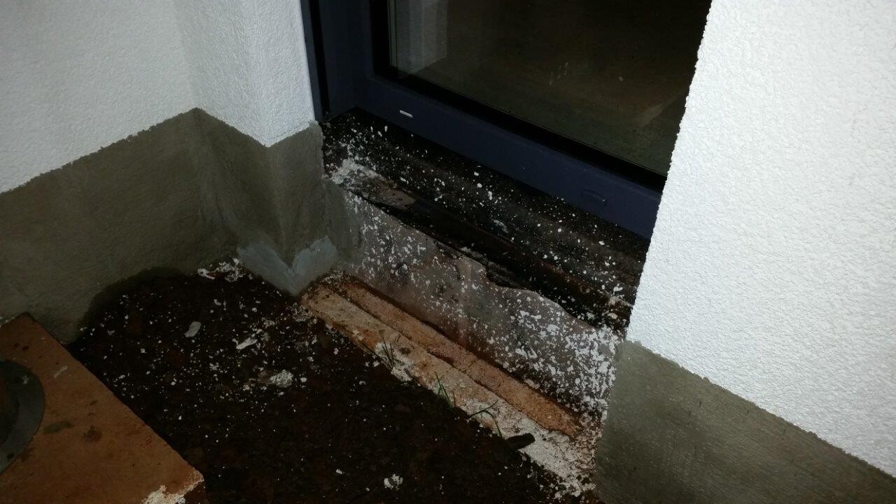 Kellerfenster im Erdreich mit Steinen und Putzresten vor weißer Außenwand.