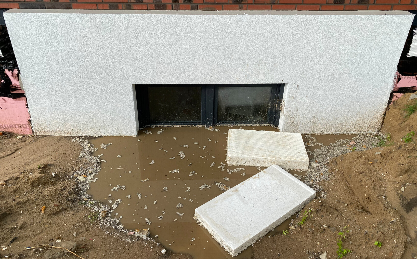 Kellerfenster in weißer, rauer Putzfassade; davor eine Schlamm-Pfütze und zwei lose Betonplatten.