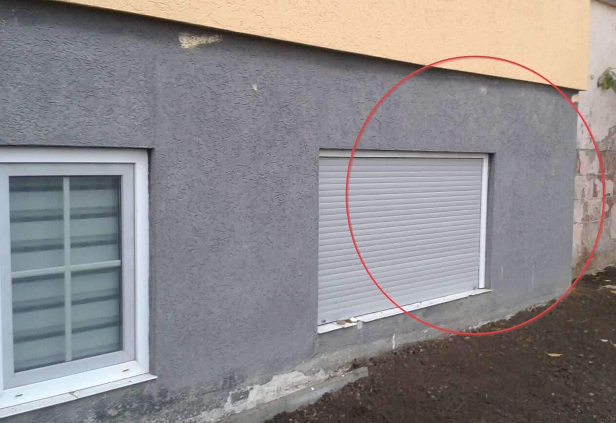 Außenansicht eines grauen Kellerbereichs mit geschlossenem Rollladenfenster