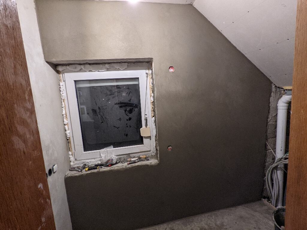 Kleines Fenster in einer unfertigen Wand; Werkzeuge liegen auf dem Fensterbrett, Rohre rechts.