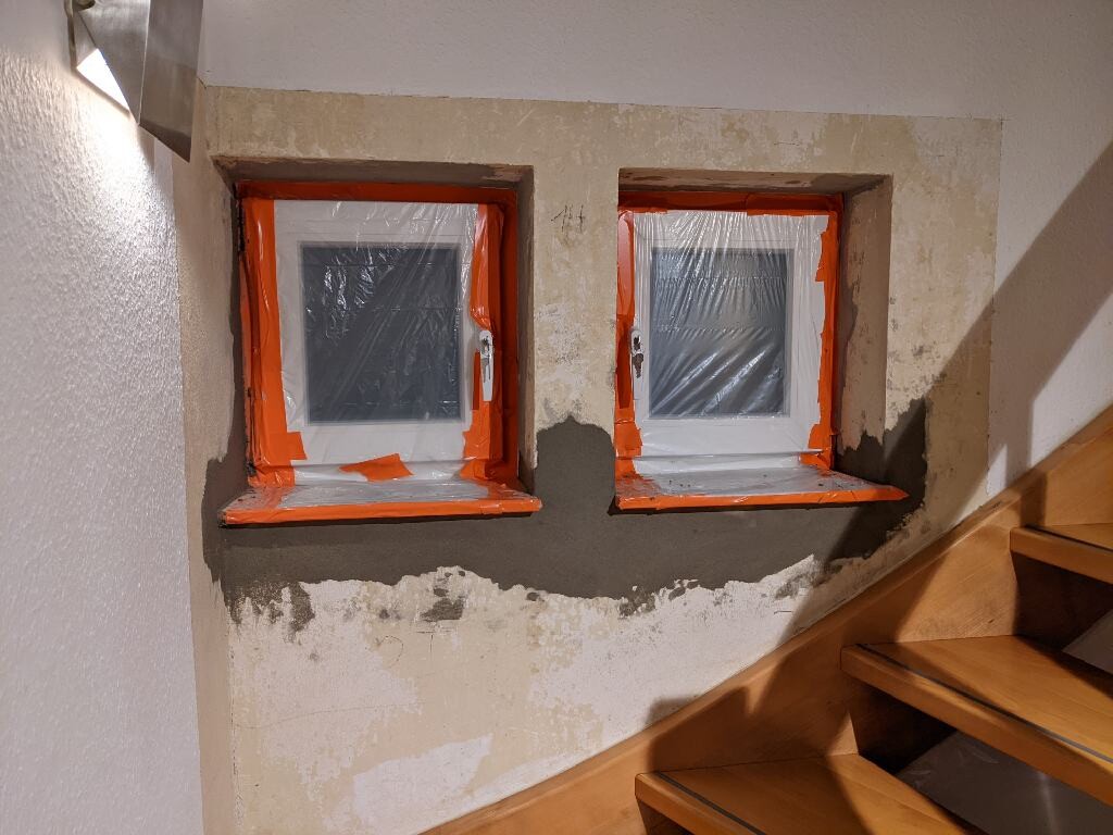 Zwei kleine quadratische Fenster in roher Wand, Plastikfolie und orangefarbenes Abdeckband.