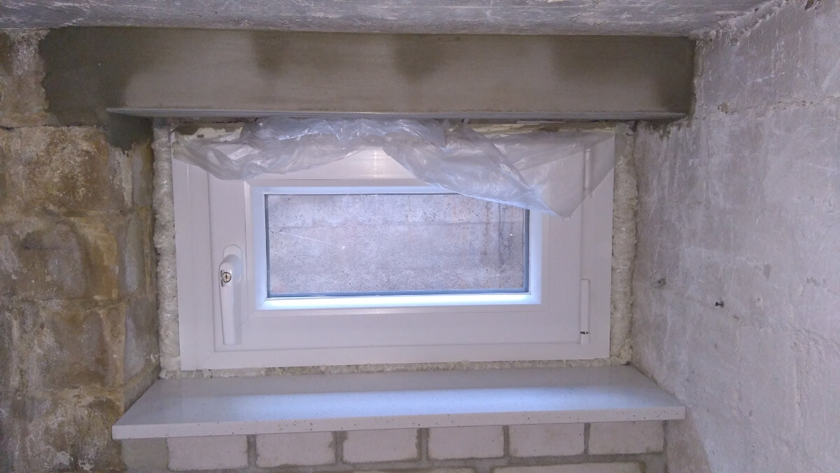 Kleines weißes Kellerfenster in Stein- und Putzwand, Plastikfolie darüber.
