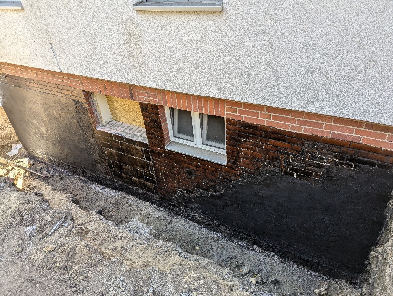 Ausgehobene Baugrube vor Kellerwand mit Fenster; dunkle Abdichtung am Fundament.