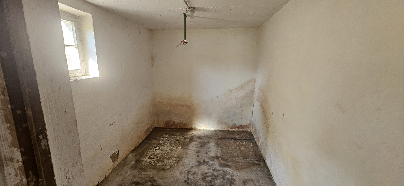 Kleines, leerstehendes Kellerzimmer mit feuchten Flecken an Boden und Wänden; linkes Fenster.