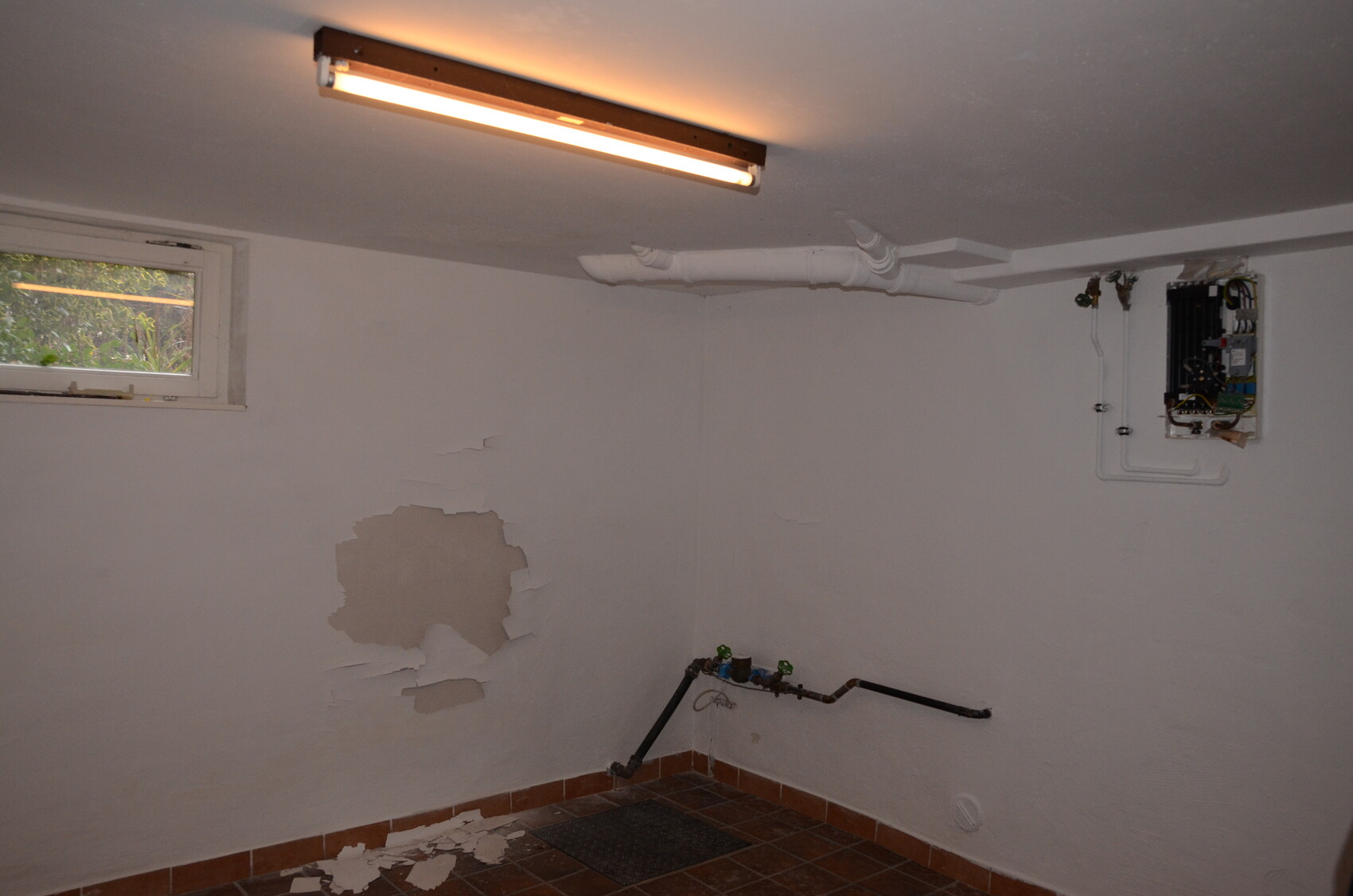 Kellerrraum mit Heizgerät an der Wand, sichtbare Rohre, beschädigte Wandfläche, Fenster links.