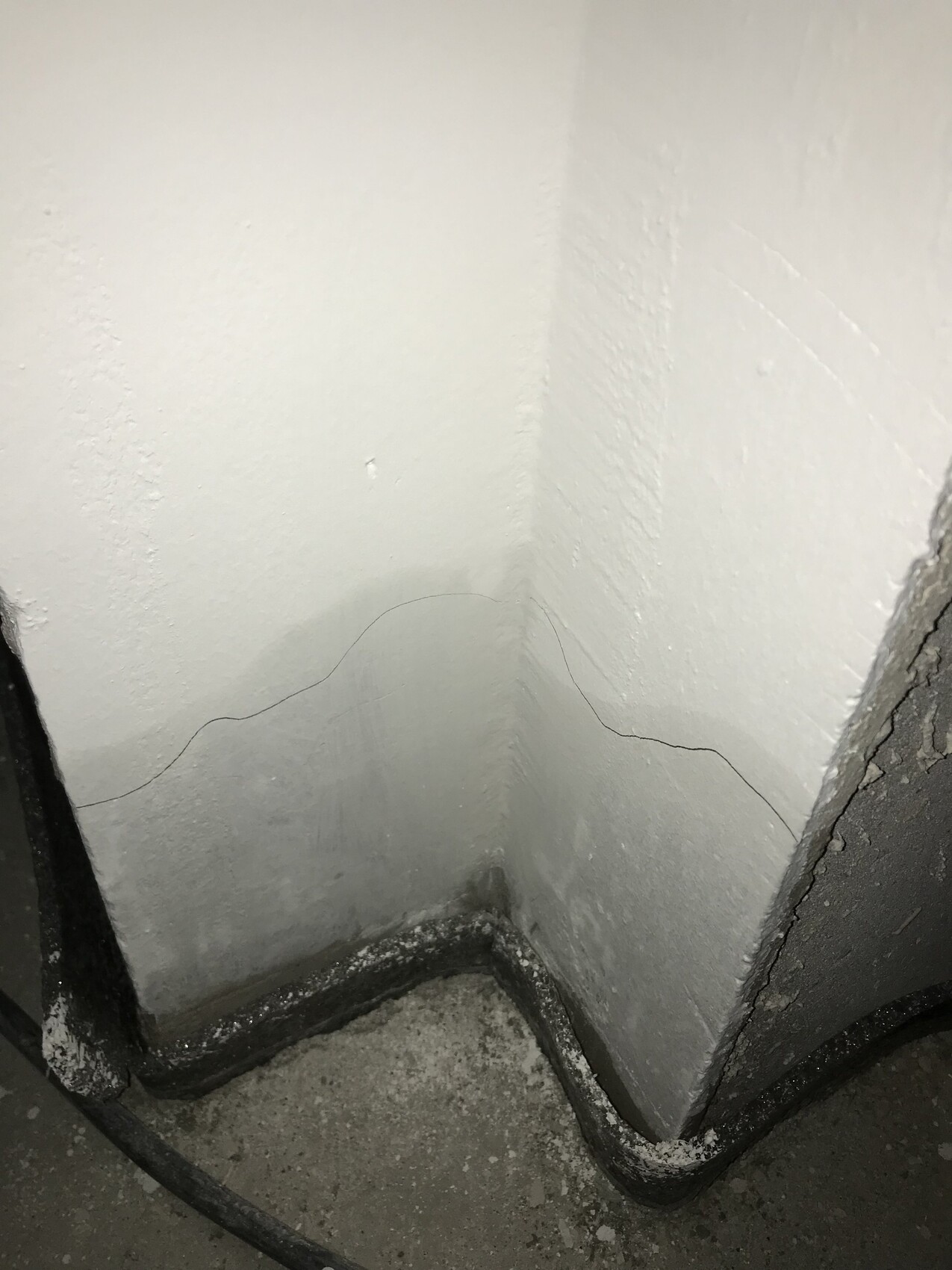 weiße Wand in der Ecke mit einer feinen, geschwungenen Linie; schwarzes Kabel am Boden.