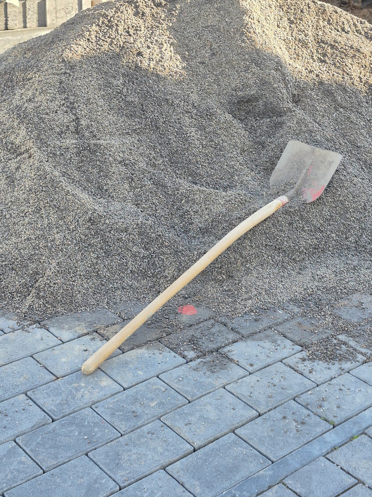Schaufel liegt schräg auf Kieshaufen neben gepflastertem Bauplatz.