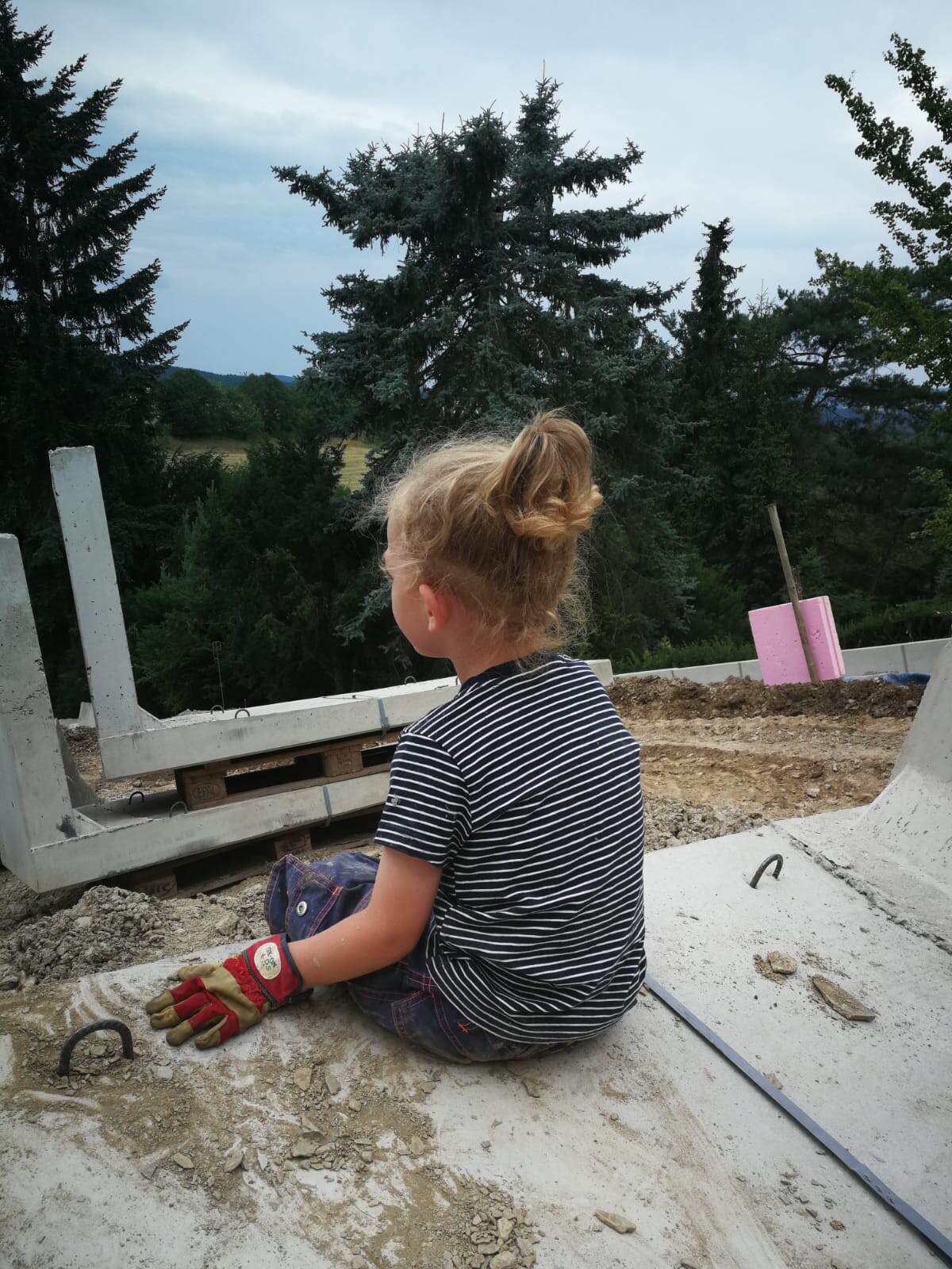 Mädchen mit Dutt sitzt auf einer Baustelle, trägt gestreiftes Shirt und Handschuhe, schaut auf Bäume.