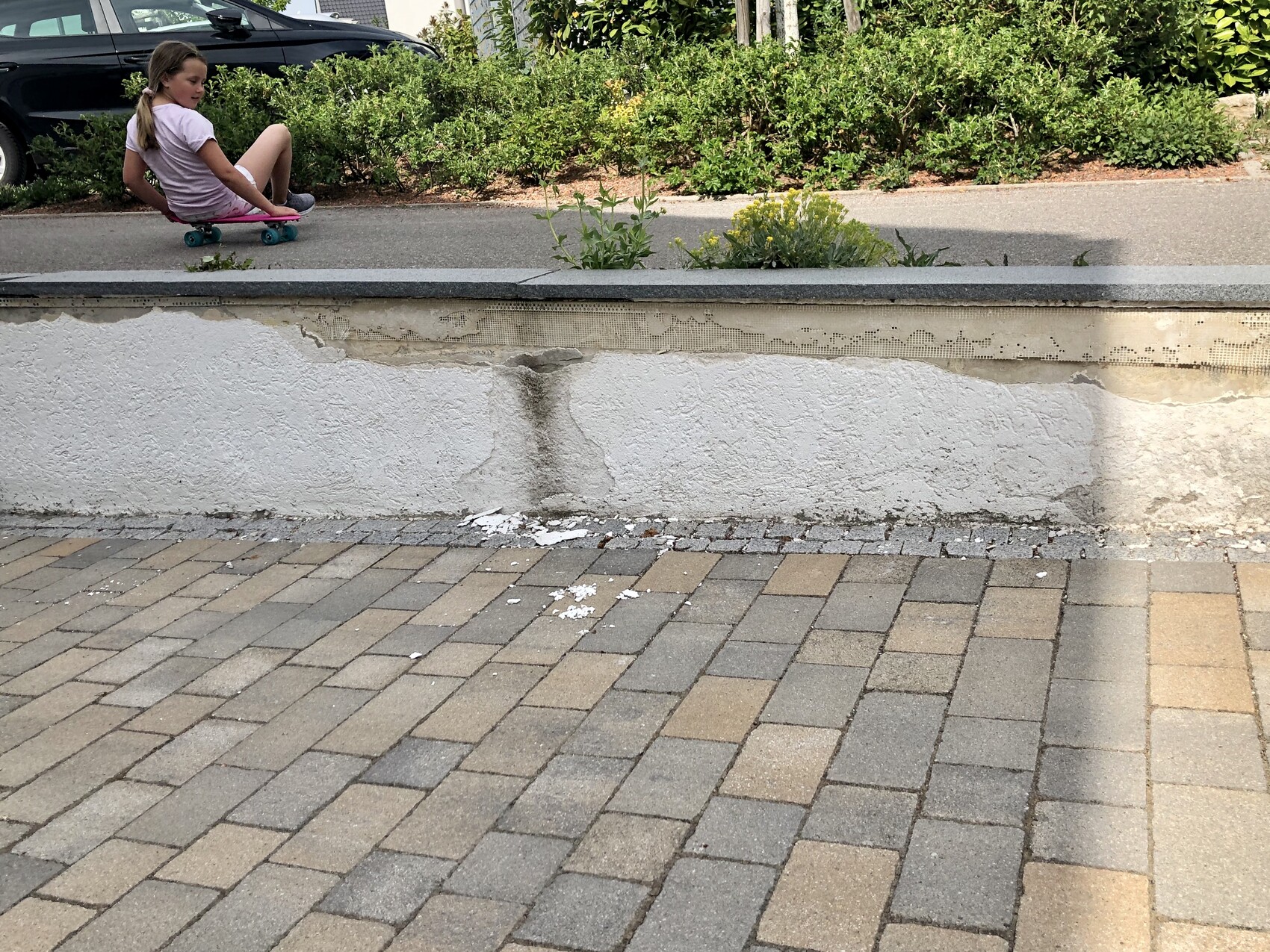 Mädchen skatet entlang eines gepflasterten Weges neben einer Mauer mit Gartenpflanzen.