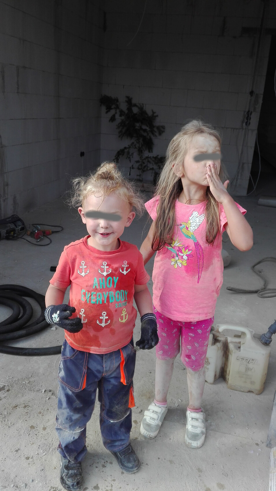Zwei verschmutzte Kinder stehen vor einer Baustelle, tragen bunte Shirts und Handschuhe.