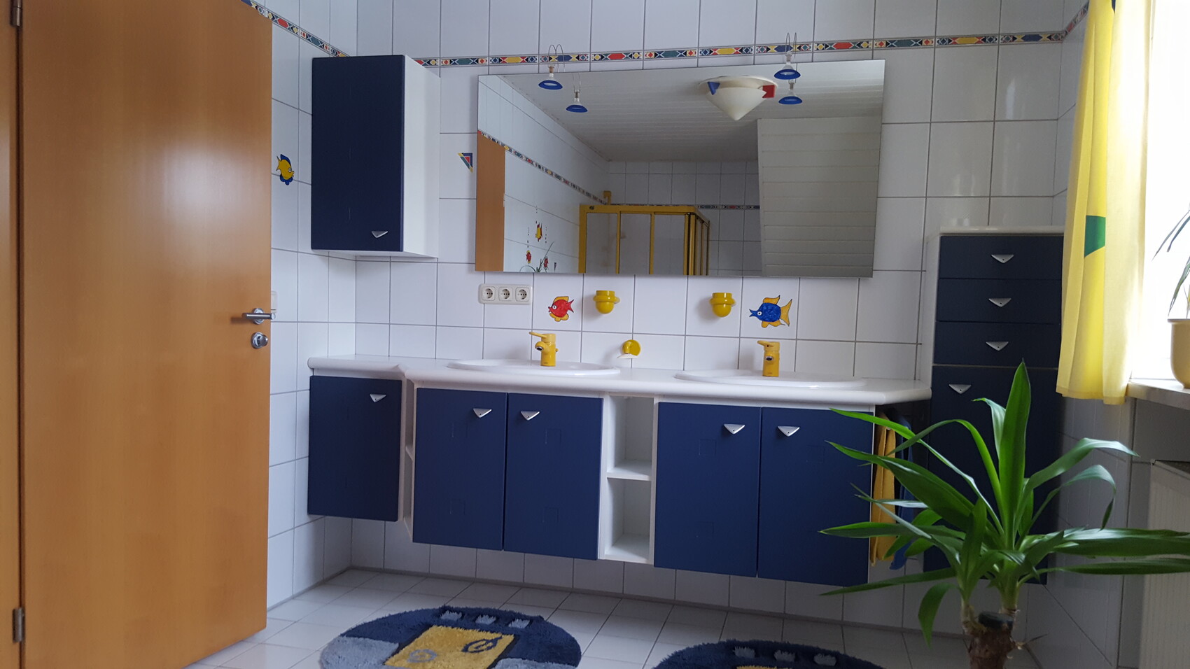 Kinderbad mit blauen Unterschränken, weißer Fliesenwand, Spiegel und gelben Armaturen sowie Pflanze
