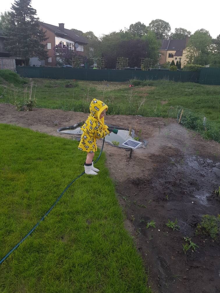 Ein Kind in gelber Minion-Jacke gießt Wasser mit Schlauch in den Garten, Häuser im Hintergrund.