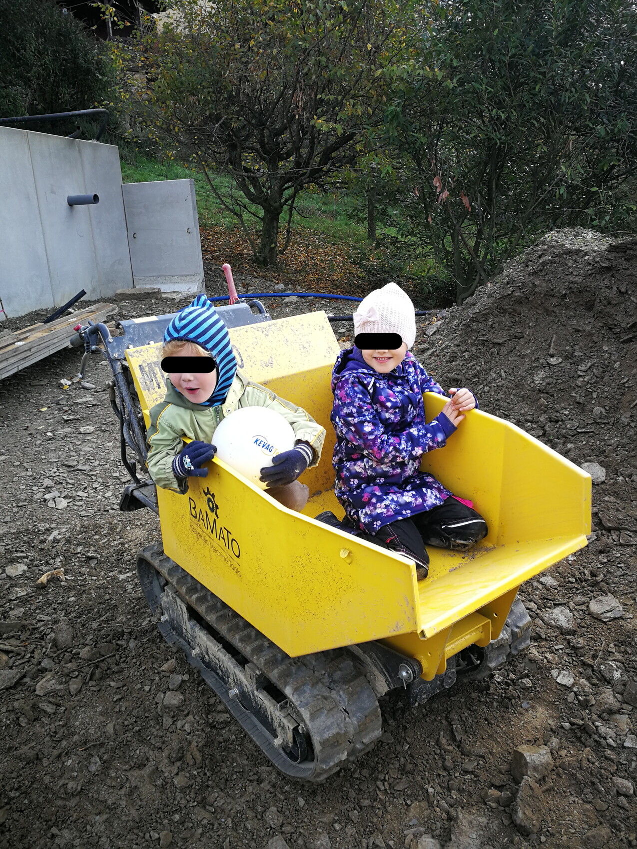Zwei Kinder sitzen in einem gelben Baukipper auf einer Baustelle, einer hält einen Helm.