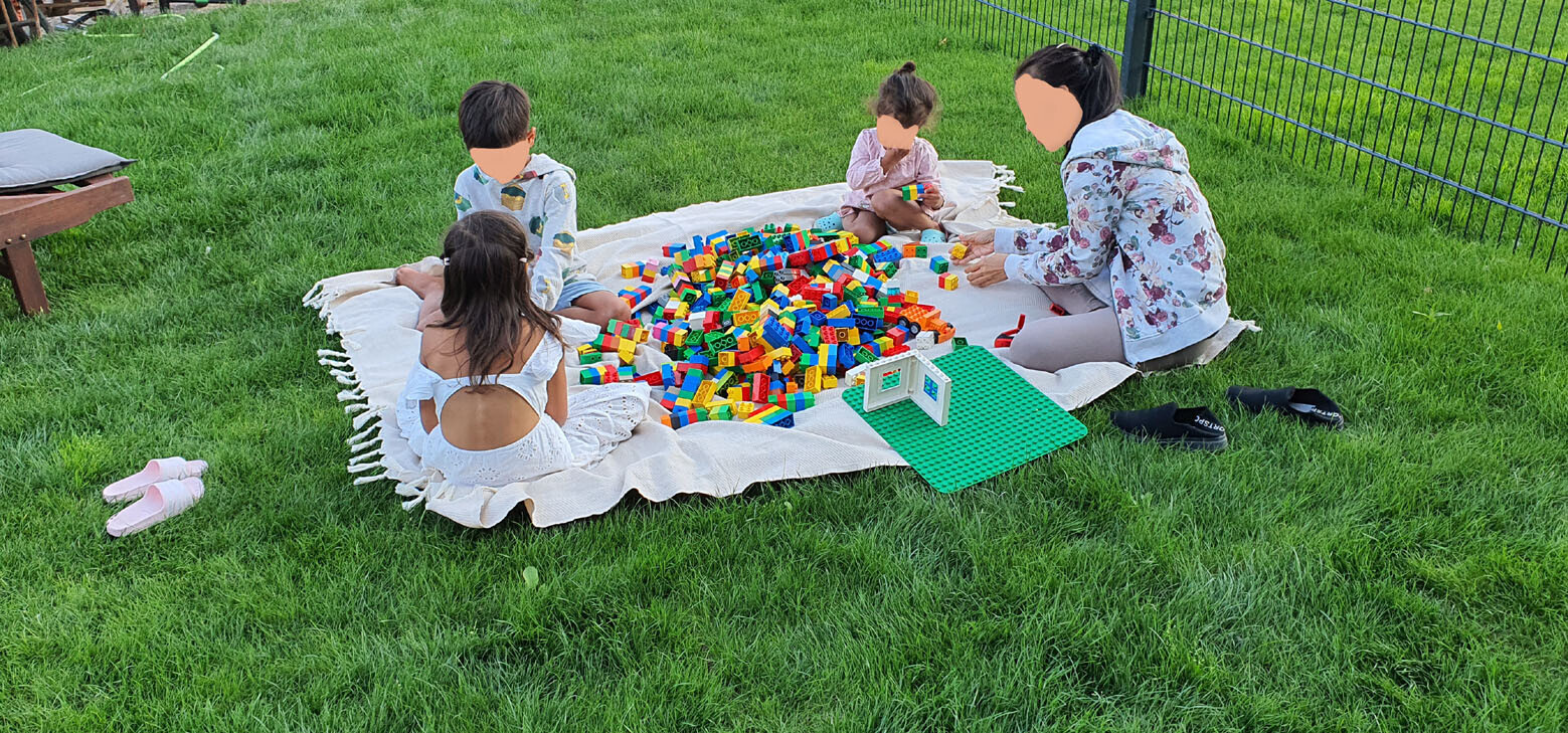 Vier Kinder spielen im Garten auf einer Decke mit bunten LEGO-Steinen und grüner Basisplatte.