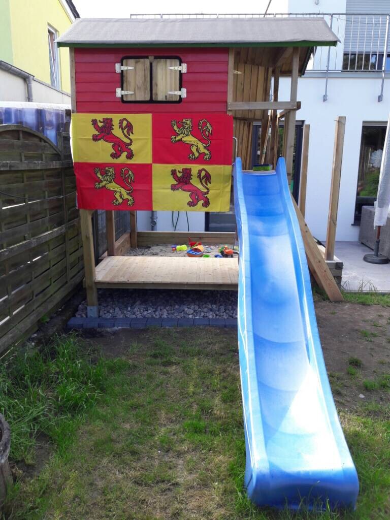 Kleines Spielhaus mit blauer Rutsche im Garten, bunte Flaggen an der Front.