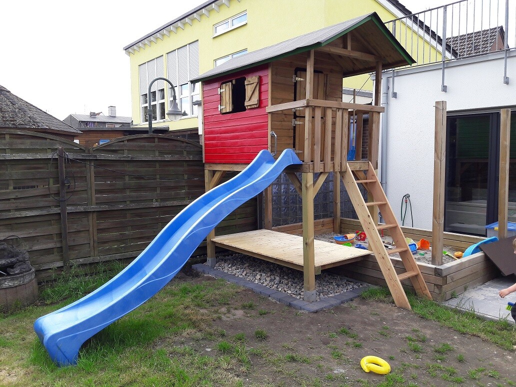 Holz-Spielturm auf Stelzen mit roter Hütte, blauer Rutsche, Leiter und Sandkasten im Garten.