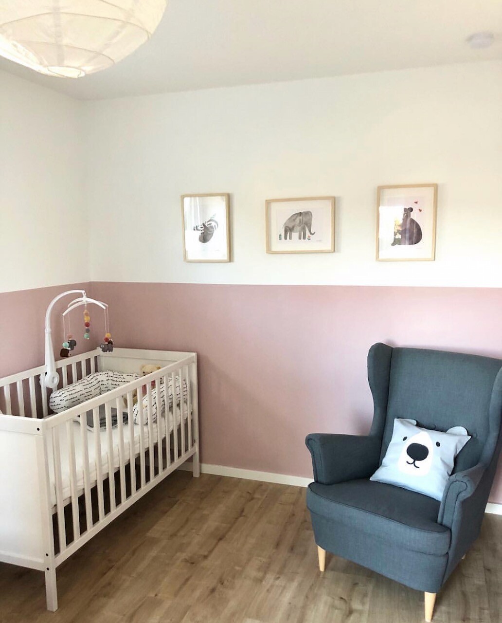 Kinderzimmer: Weißes Babybett mit Mobile, rosa Wand, blauer Sessel mit Bärenkissen, Tierbilder.