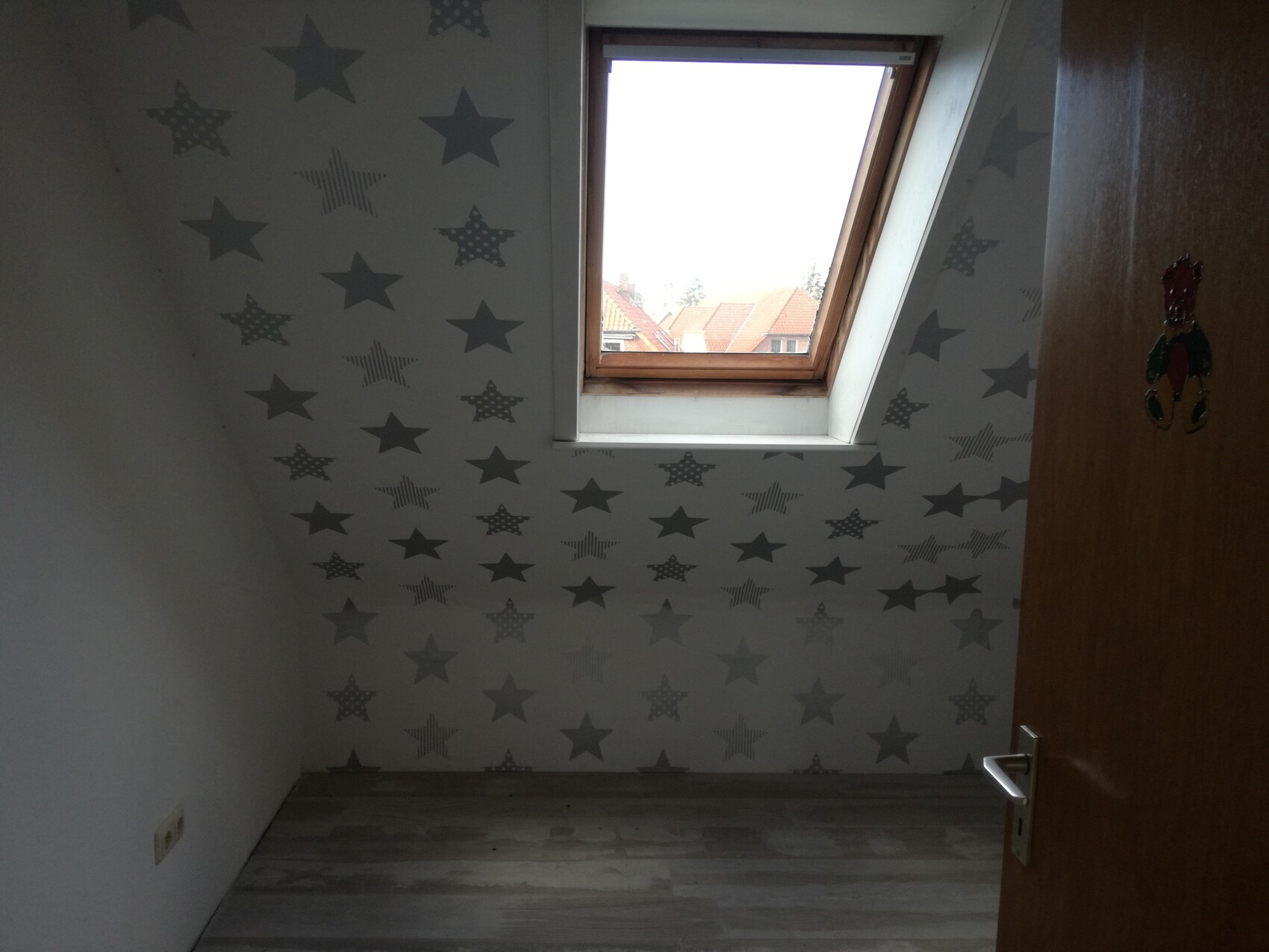 Kleines Dachbodenzimmer mit Sternen-Wandtapete, Dachfenster und Holzboden; Tür rechts.