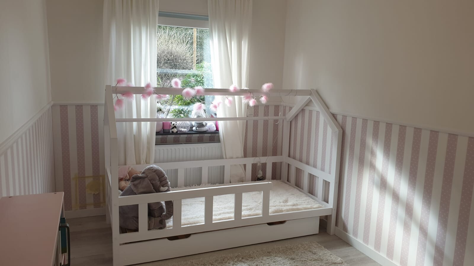 Weißes Hausbett mit rosa Pompon-Girlande, Pastellstreifen-Wand und Kuscheltiere am Fenster.