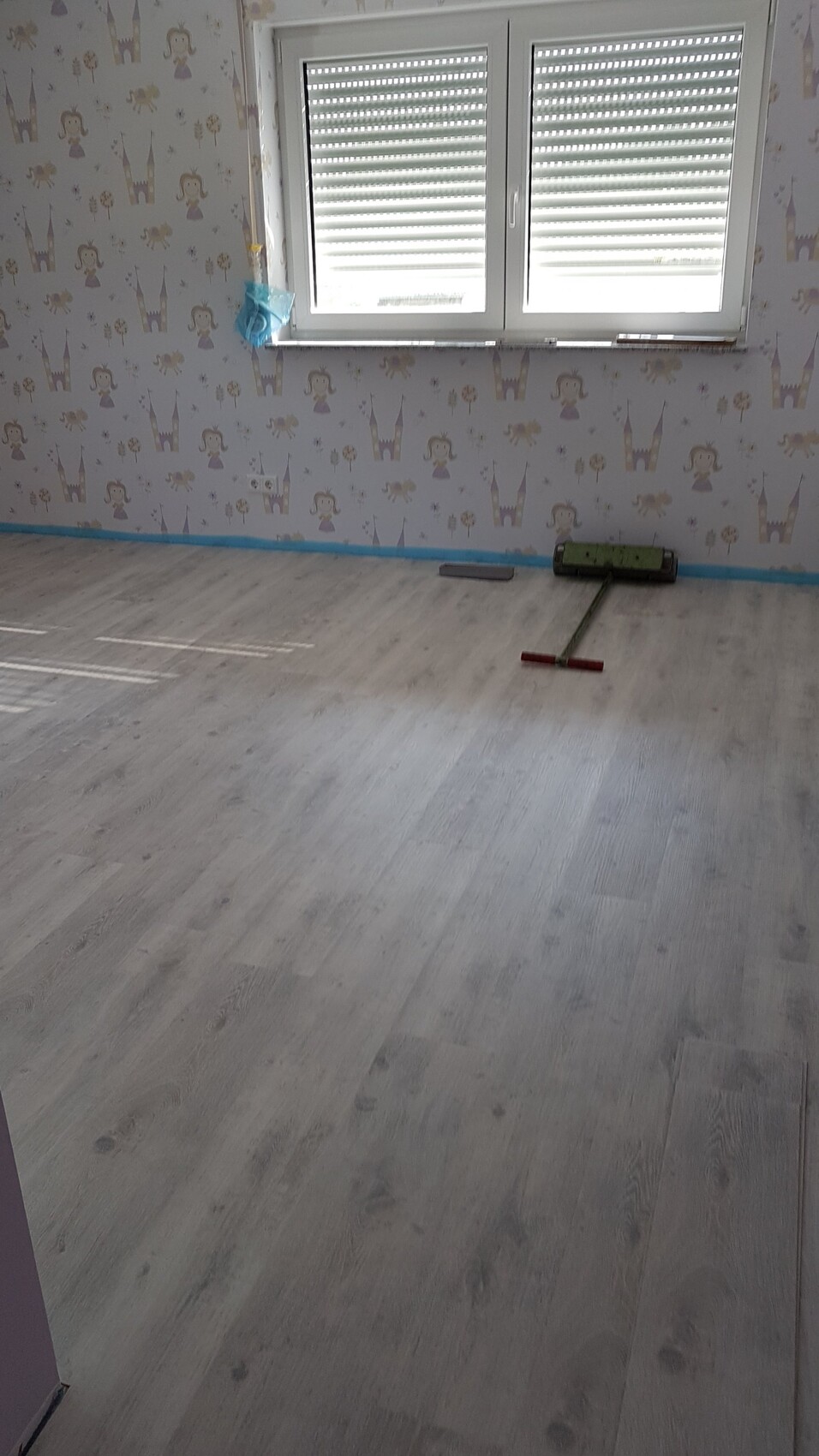 Leeres Kinderzimmer in Renovierung: grauer Laminatboden, Prinzessinnen-Tapete, Fenster mit Rollläden.