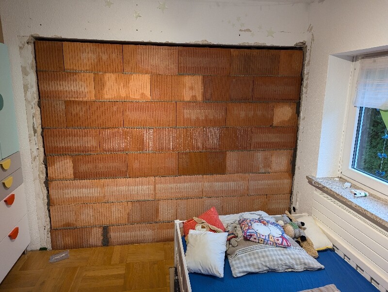 Kleines Zimmer mit freigelegter Backsteinwand, Bett mit Kissen und Stofftiere, Fenster rechts.