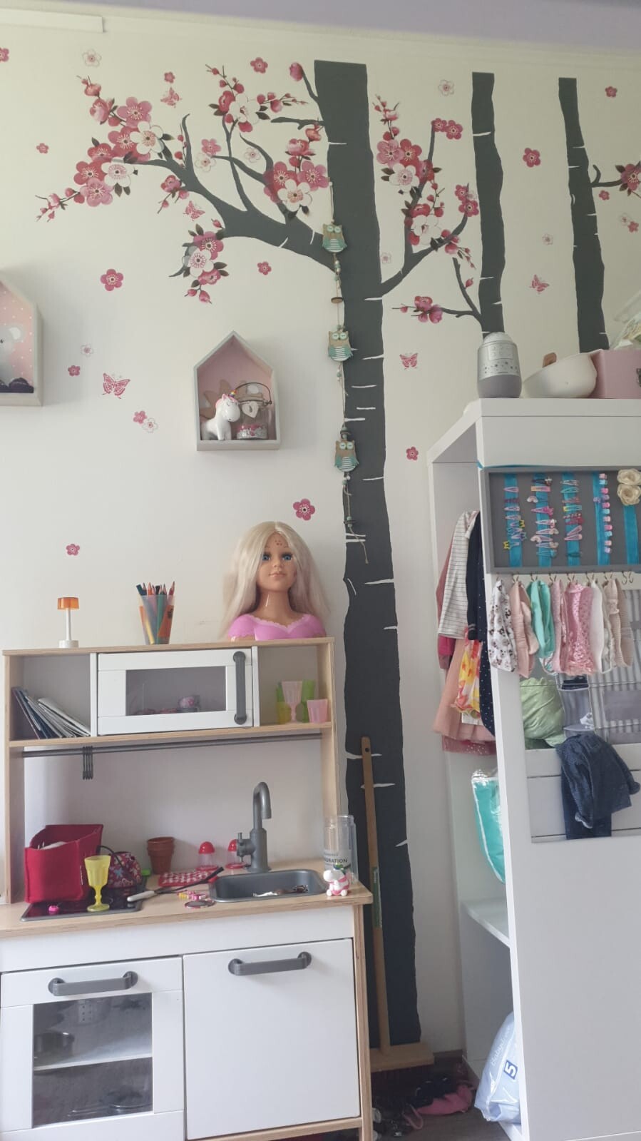 Kinderzimmer mit Kirschblüten-Wandbild, Puppenkopf auf Spielküche, Kleiderschrank rechts.