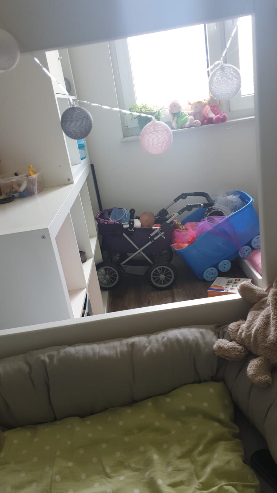 Kinderzimmer mit Puppenwagen, Spielzeugkiste und Kuscheltieren am Fenster, Bett im Vordergrund.