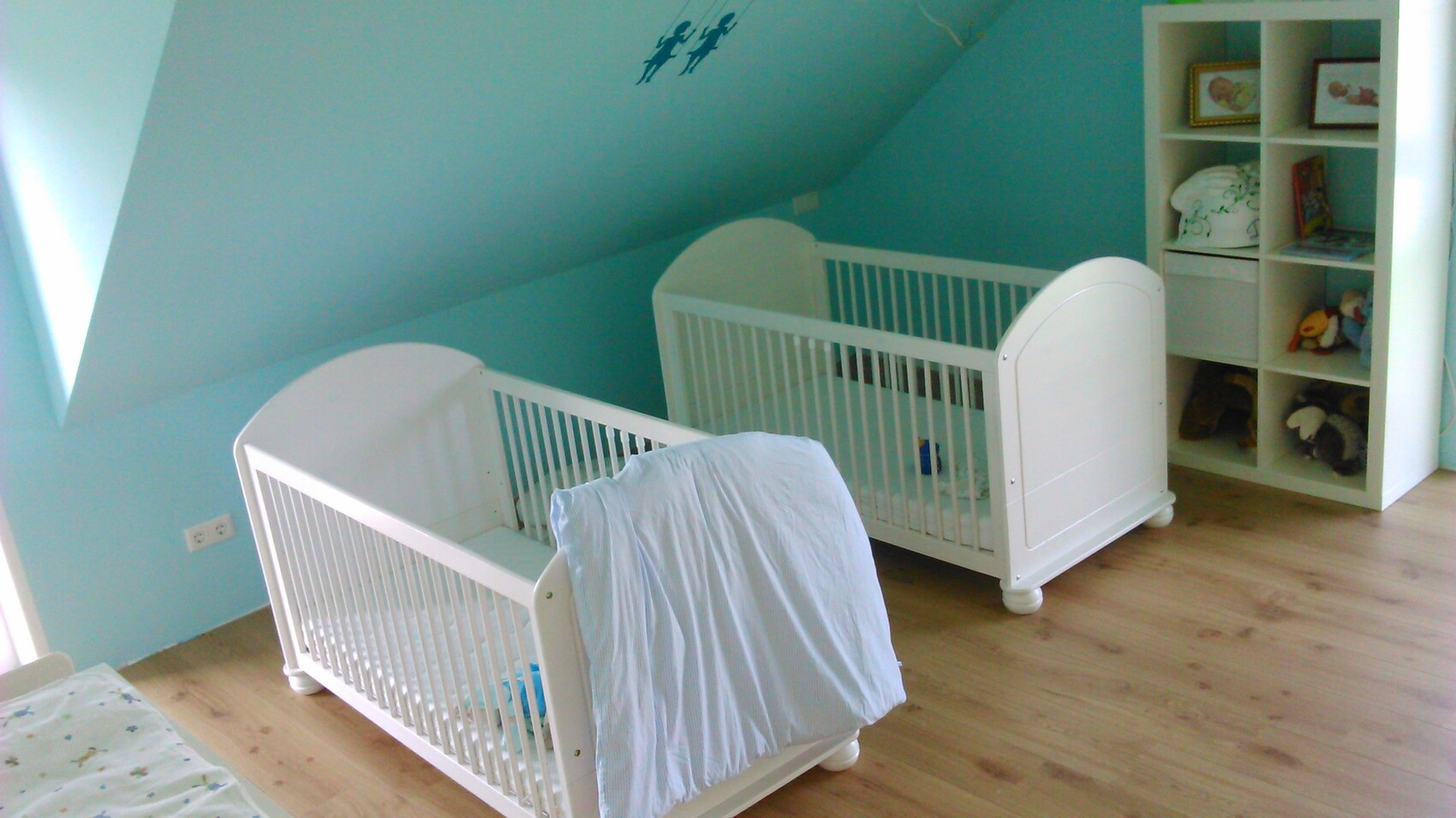 Zwei weiße Babybetten in türkisfarbenem Kinderzimmer, Regal mit Stofftieren und Holzboden.