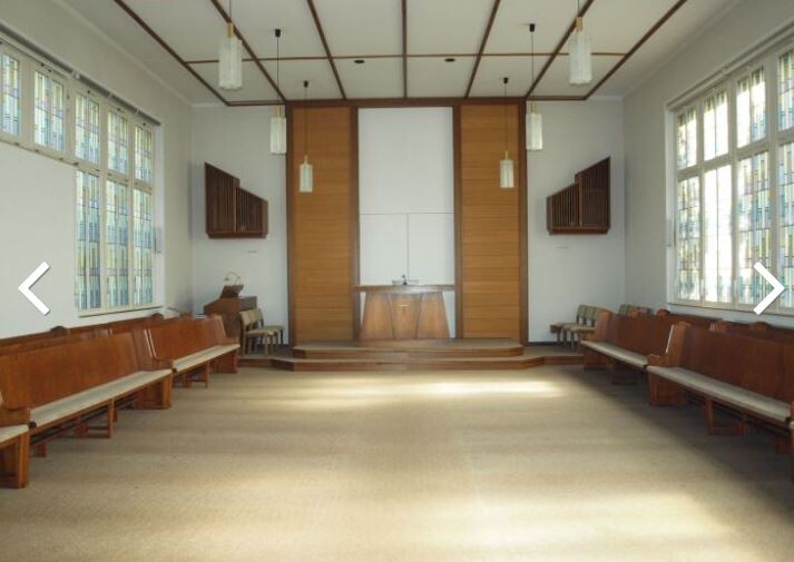 Innenraum einer Kirche mit Holzbänken, Altar und großen Fenstern.