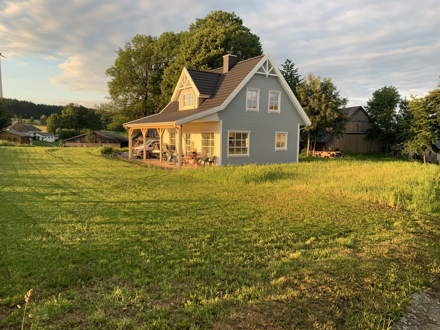 Hellblaues Einfamilienhaus mit Veranda, Auto darunter und Gartenmöbeln im Abendlicht.