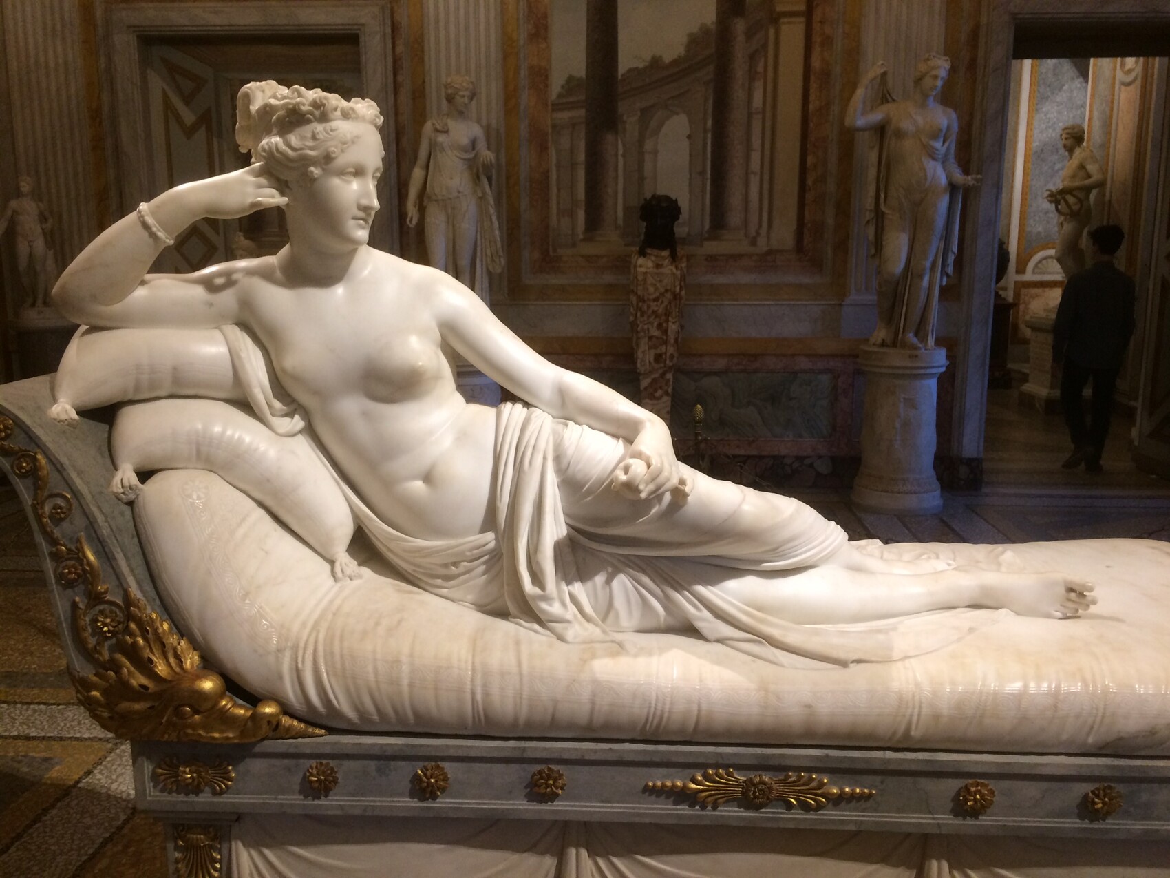 Liegende weiße Marmorskulptur einer Frau auf prunkvoller Liege im Museum.