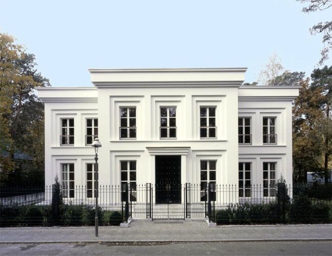 Weiße, zweistöckige Villa mit symmetrischer Fassade, zentralem Eingang und Gartenzäunen vor der Straße.