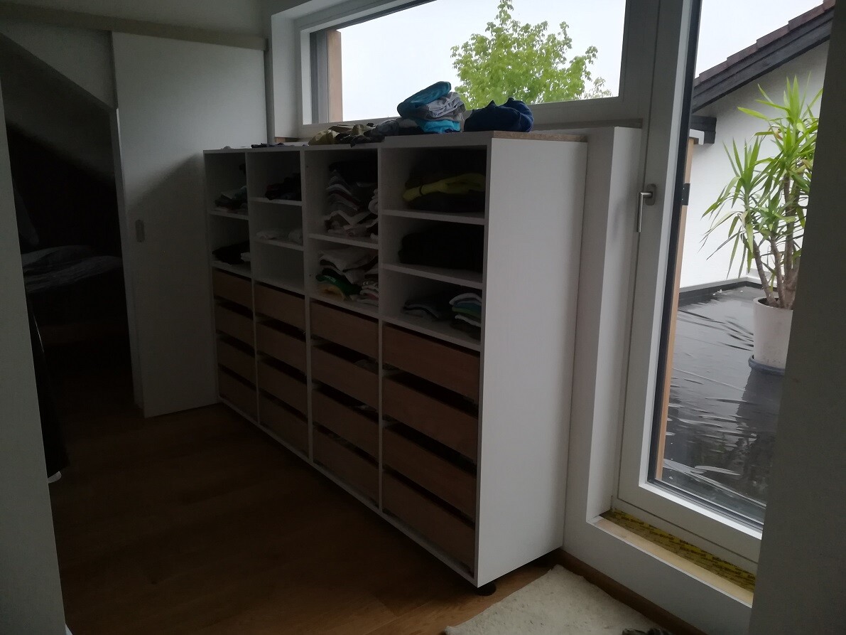 Weißes Regalsystem mit offenen Fächern voller Kleidung, neben Fenster.