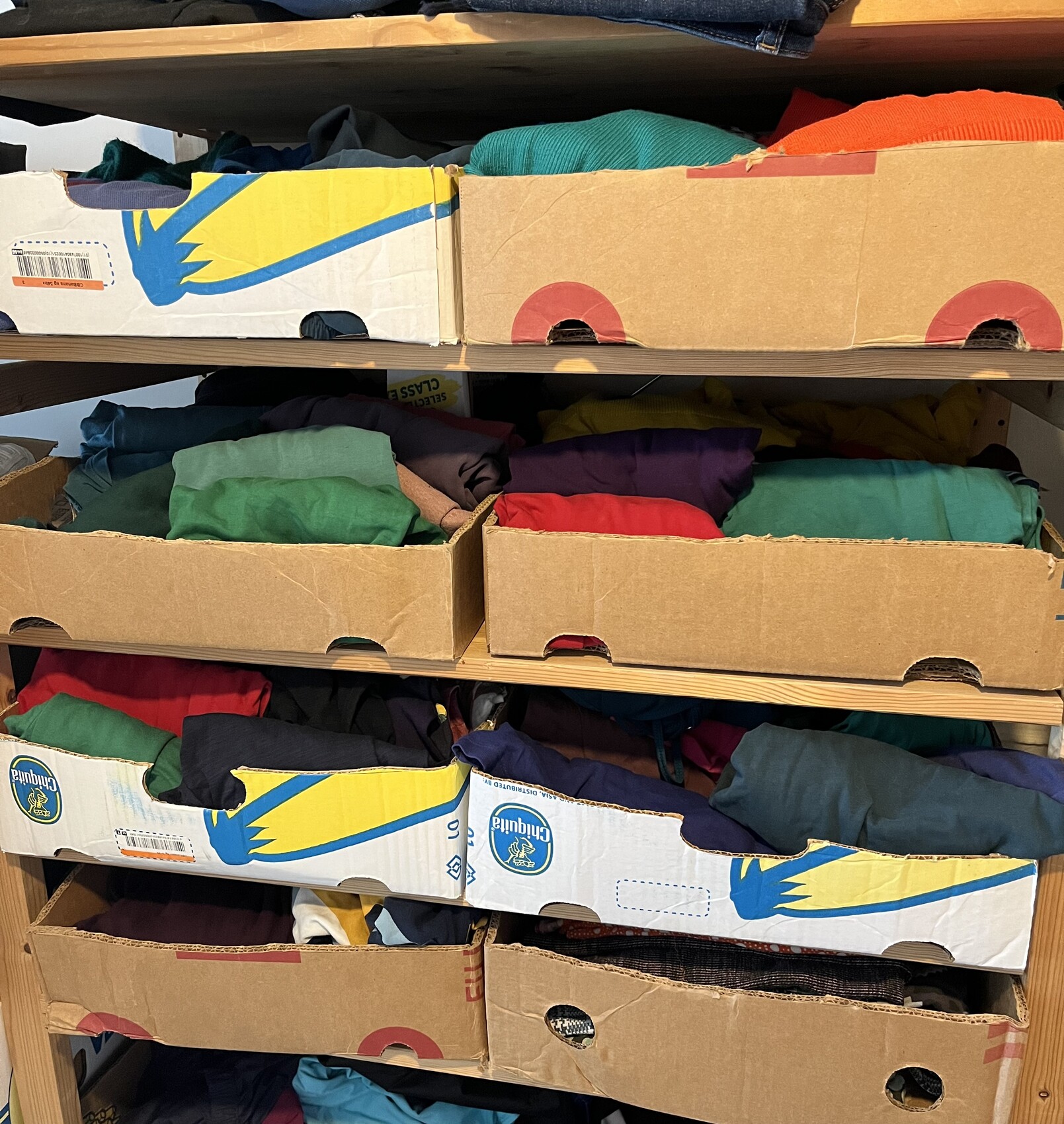 Bunte Kleidung in Kartons auf einem Holzregal, verschiedenfarbige Stoffe gestapelt.