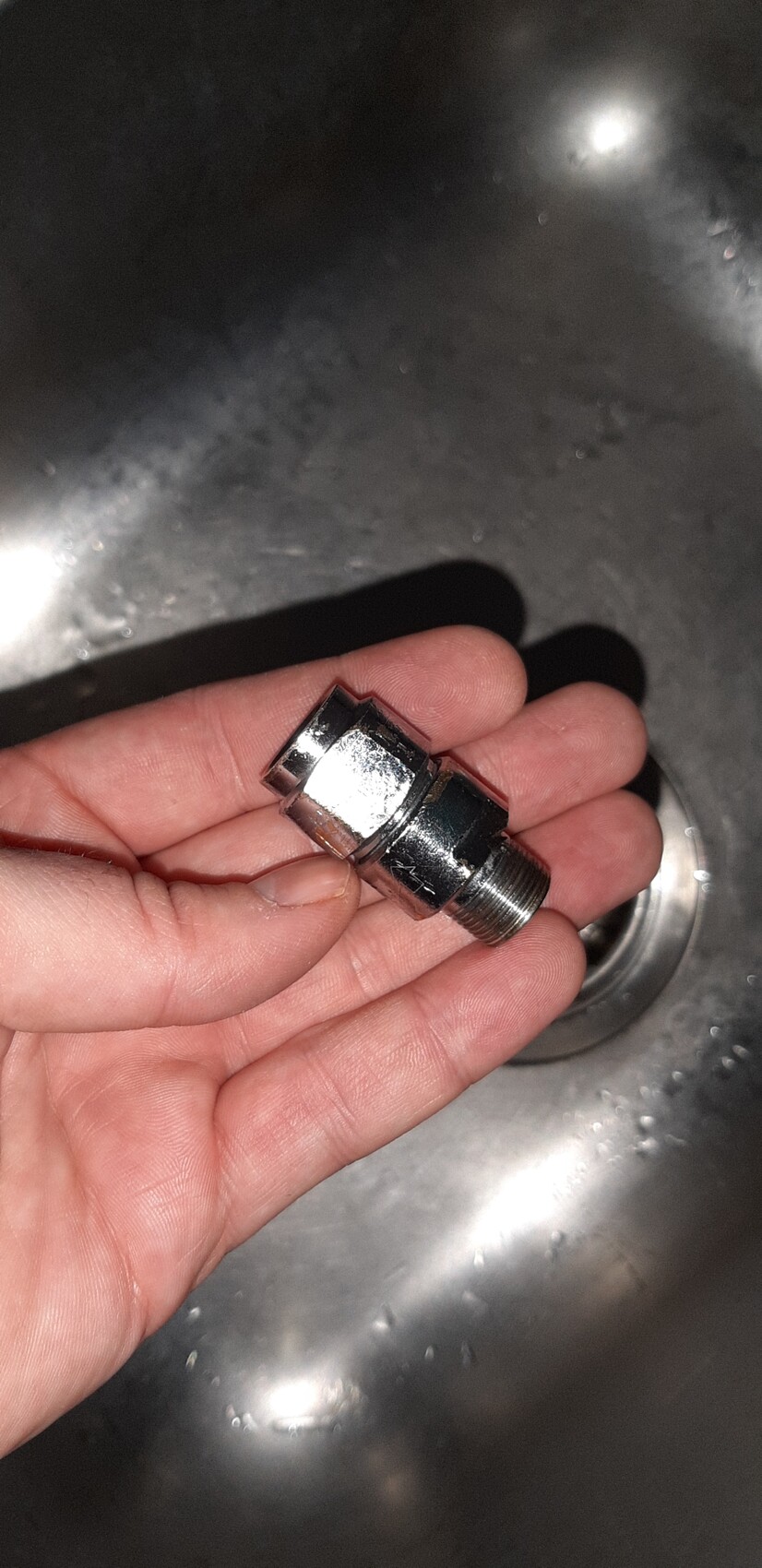 Kleines chromfarbenes Metallteil mit Gewinde, in der Hand; Spüle im Hintergrund.