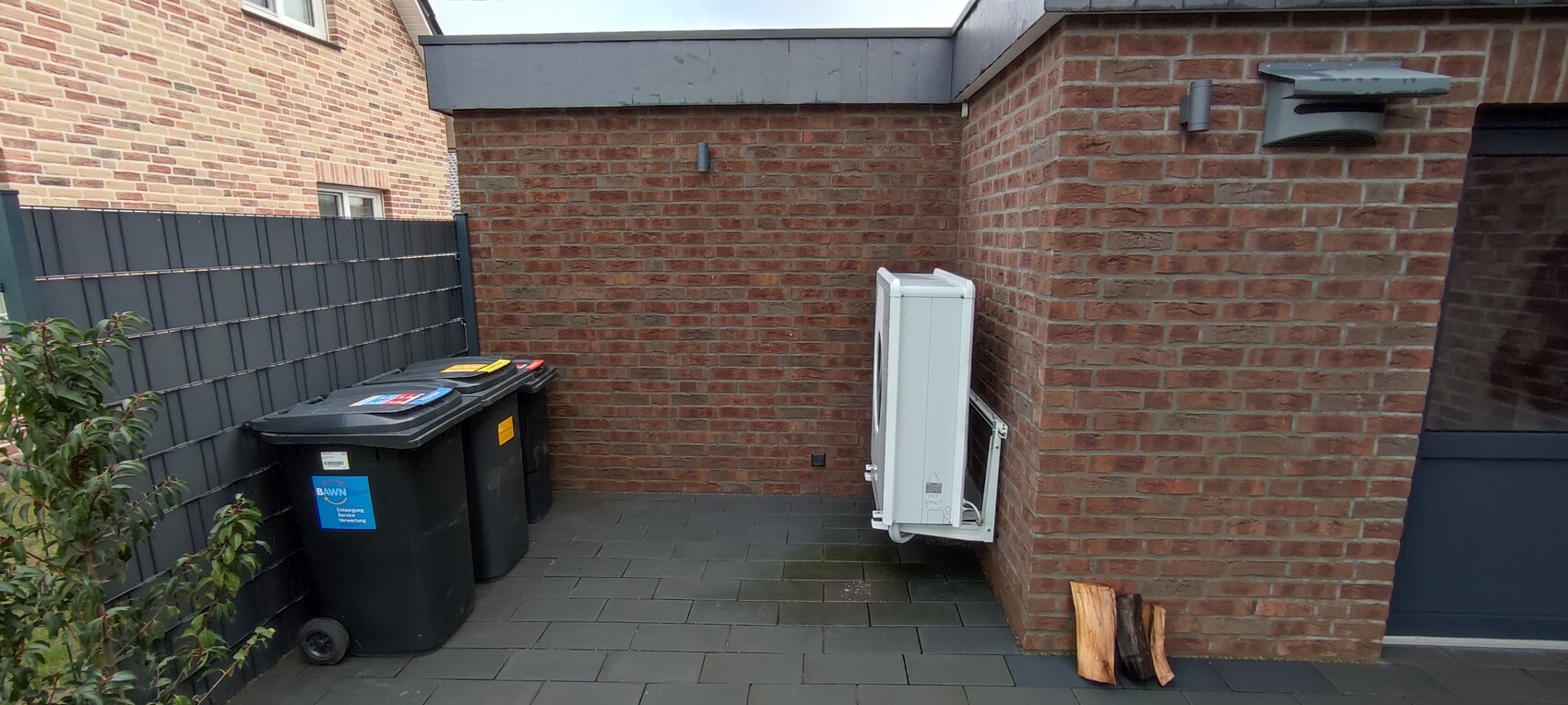 Hinterhof mit Mülltonnen links, weiße Außen-Klimaanlage an Ziegelwand rechts, Holzscheite am Boden.