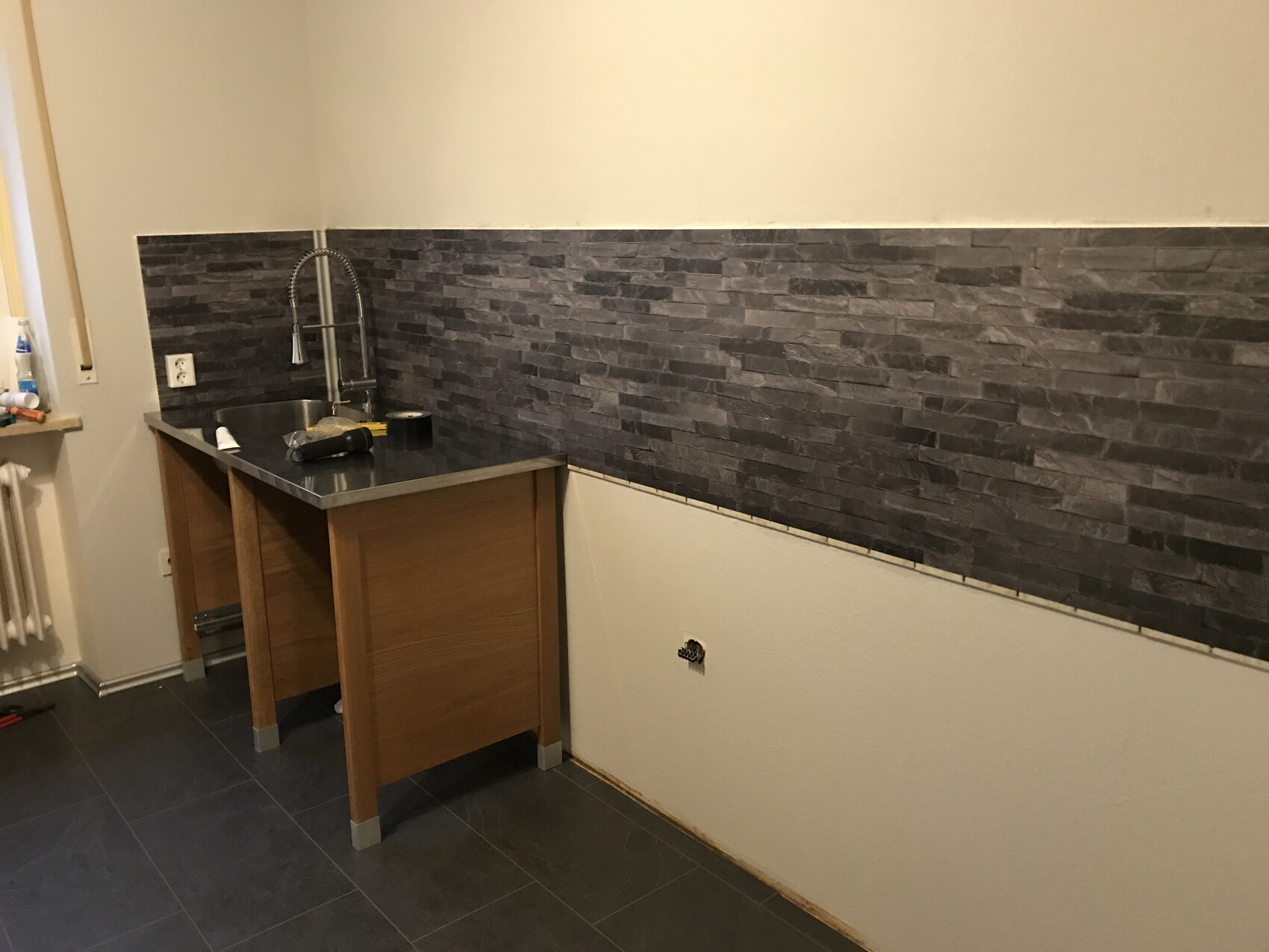 Küche mit Spüle und Armatur, dunkler Steinrückwand, Holzunterschrank und grauen Bodenfliesen.