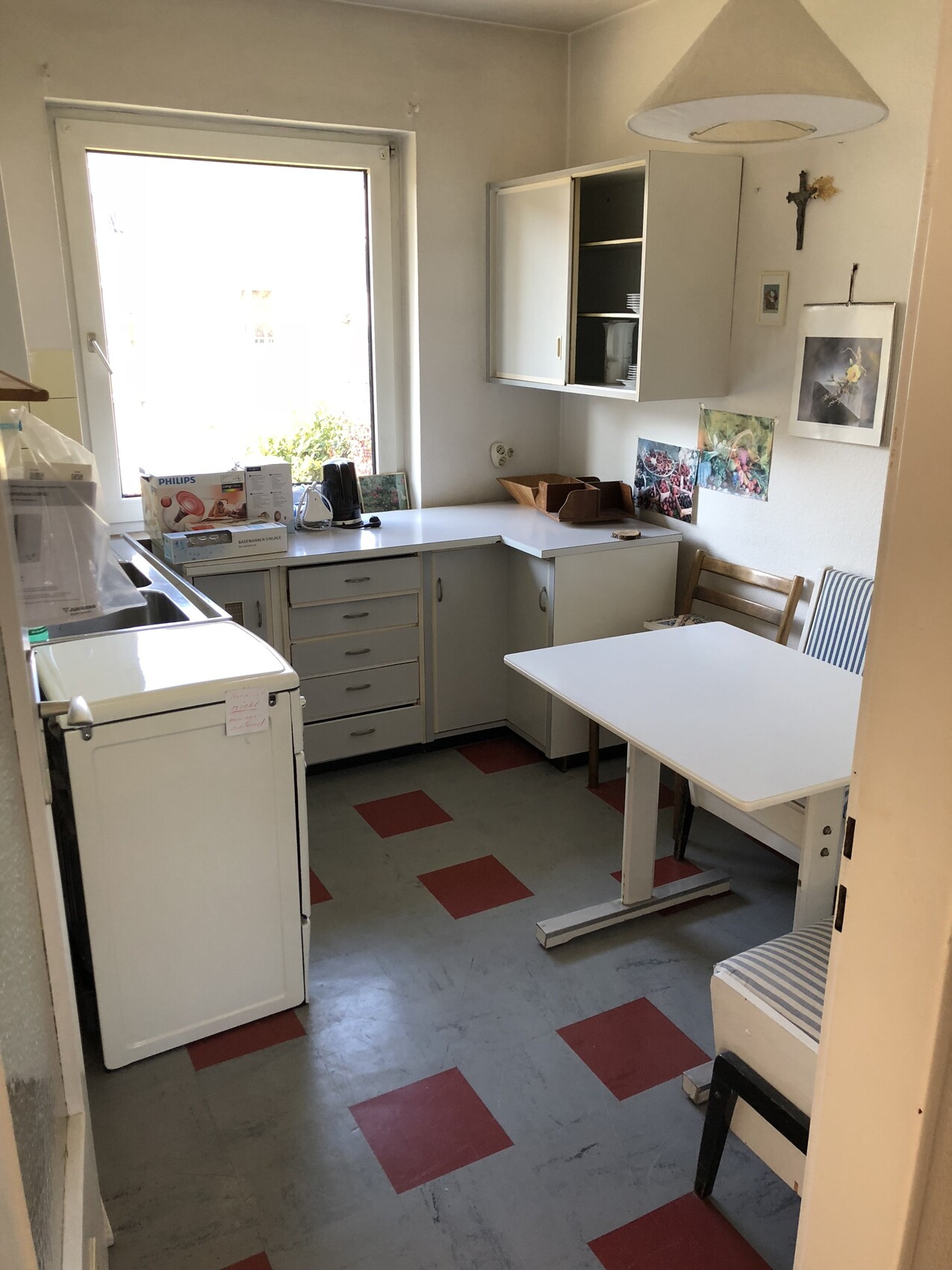 Kleine Küche mit weißer Küchenzeile, Esstisch und Stühlen, großem Fenster und Kreuz an der Wand.