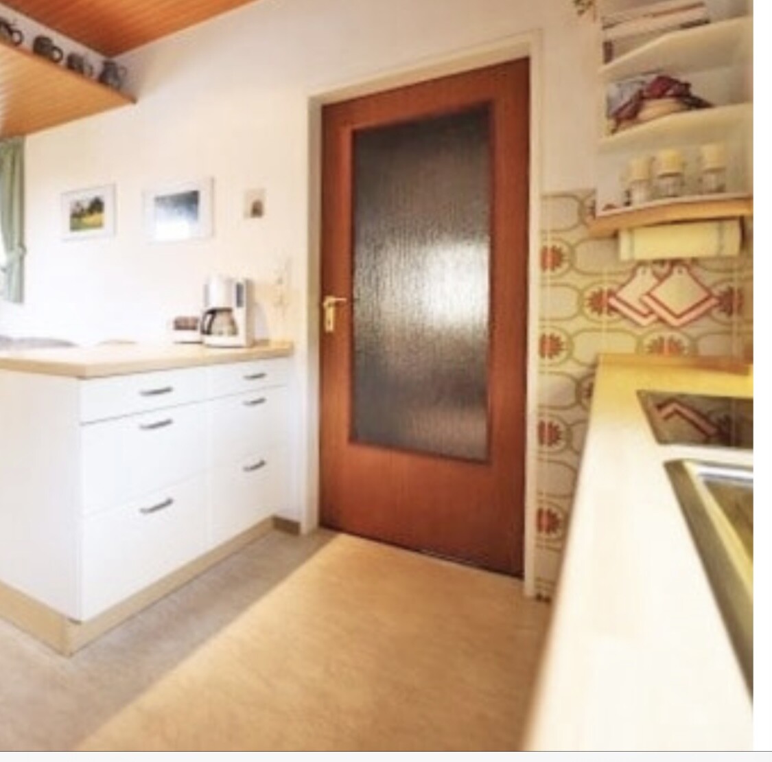 Küchenbereich mit weißen Unterschränken, Kaffeemaschine, Holztür mit Glas, Regale an Mustertapete.