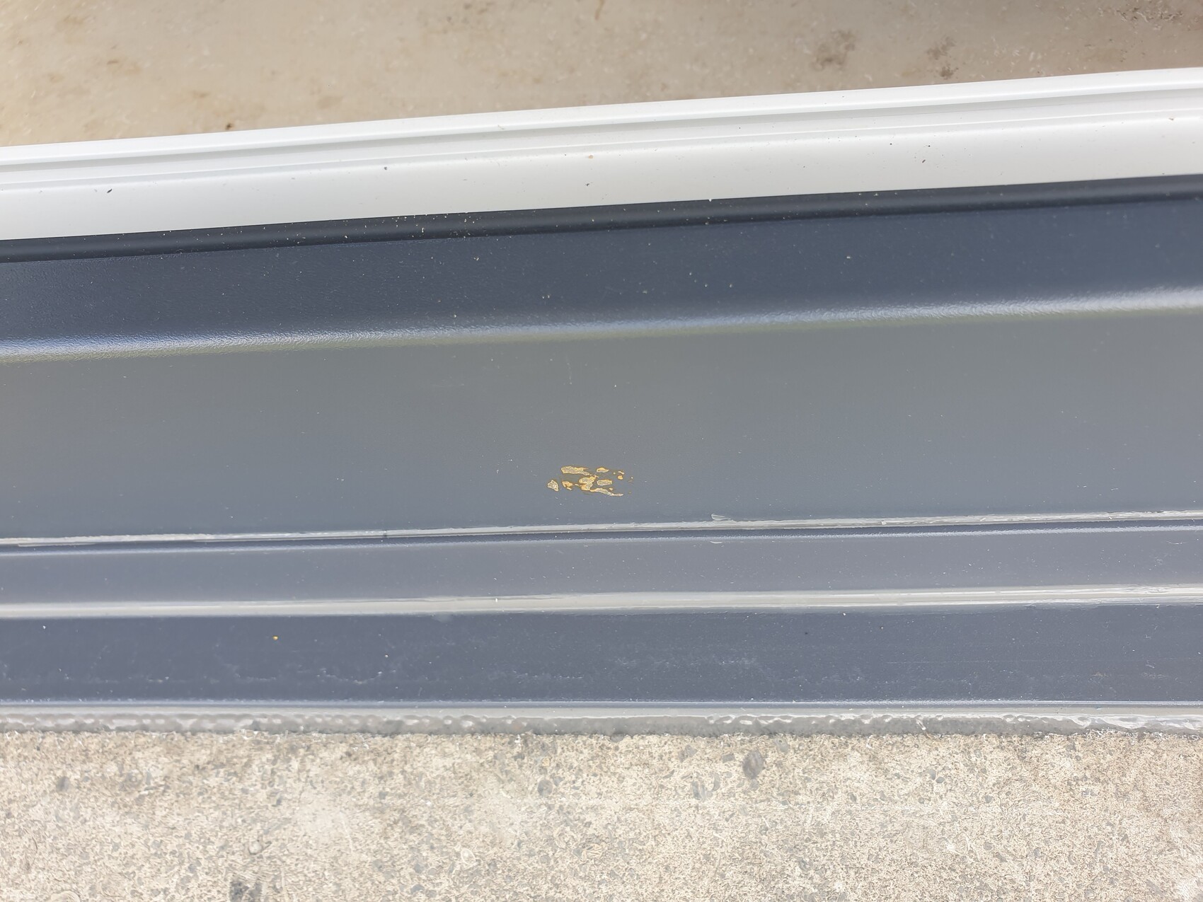 Nahaufnahme einer graublauen Metalloberfläche mit kleinem braunen Rostfleck, Betonboden unten.