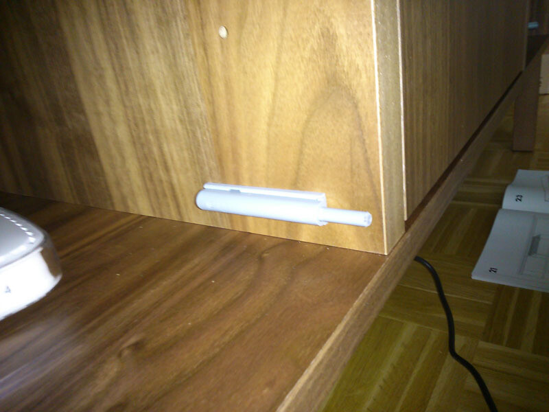 Kleiner weißer Plastikclip an der Unterseite eines Holzschranks, daneben liegt ein Kabel.