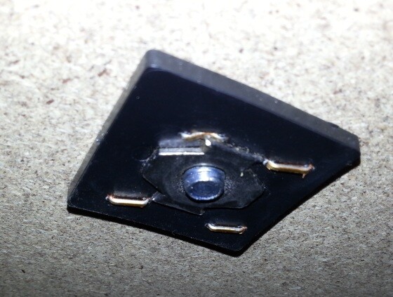 Kleine schwarze Montageplatte mit zentraler Öffnung und vier Metallpins auf rauem Betonboden.