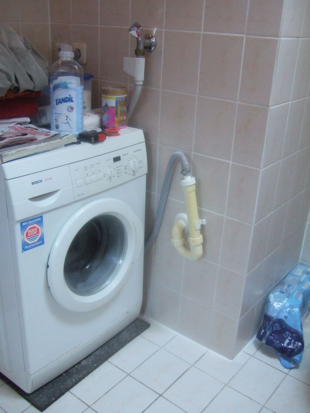 Waschkueche mit Waschmaschine, Reinigungsmittel und sichtbaren Wasserleitungen.