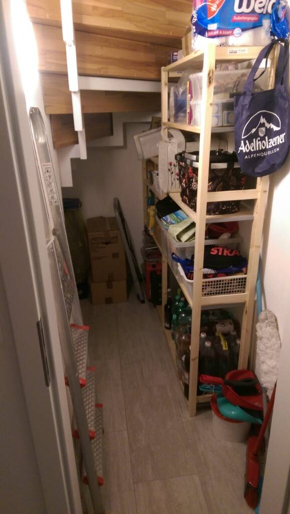 Enge Abstellkammer mit Holzregal voller Flaschen, Kisten, Reinigungsmittel, Leiter.