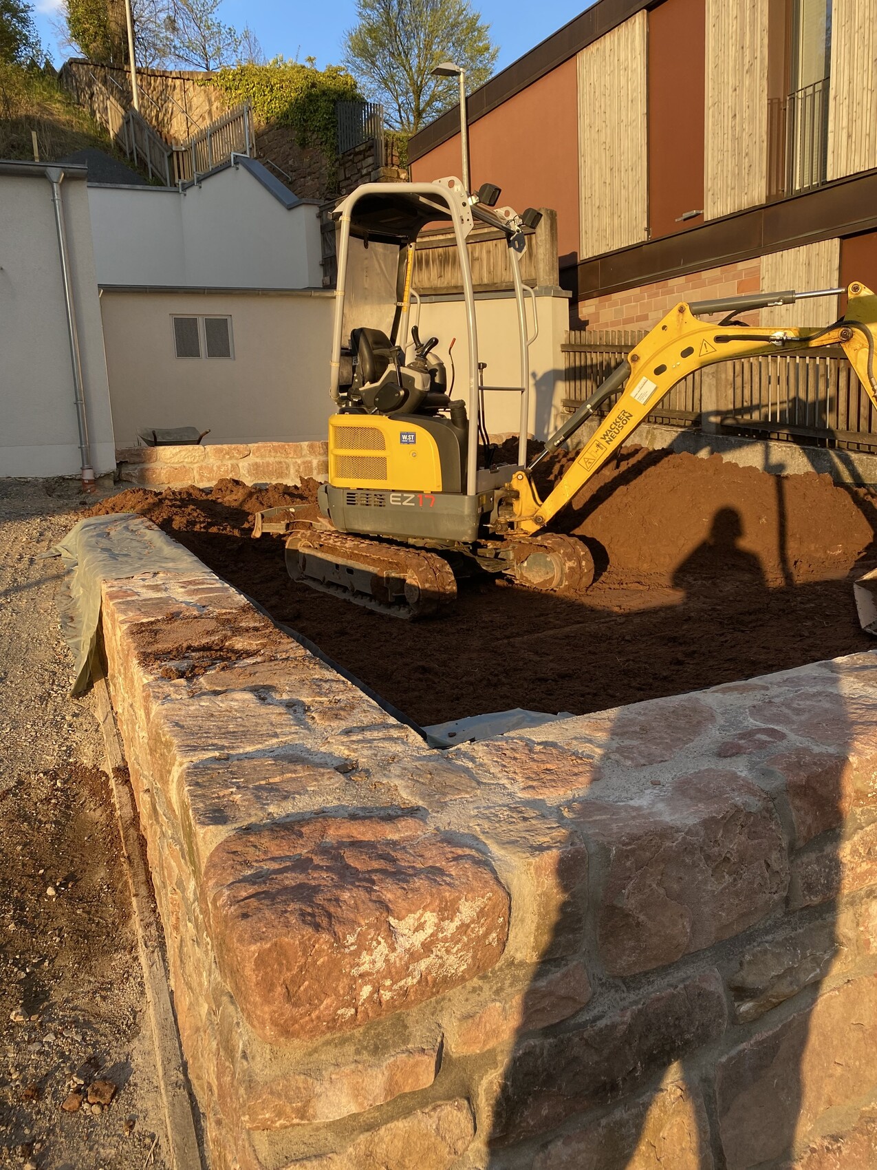 Kleiner gelber Minibagger gräbt Erde neben einer Steinmauer auf einer Baustelle.