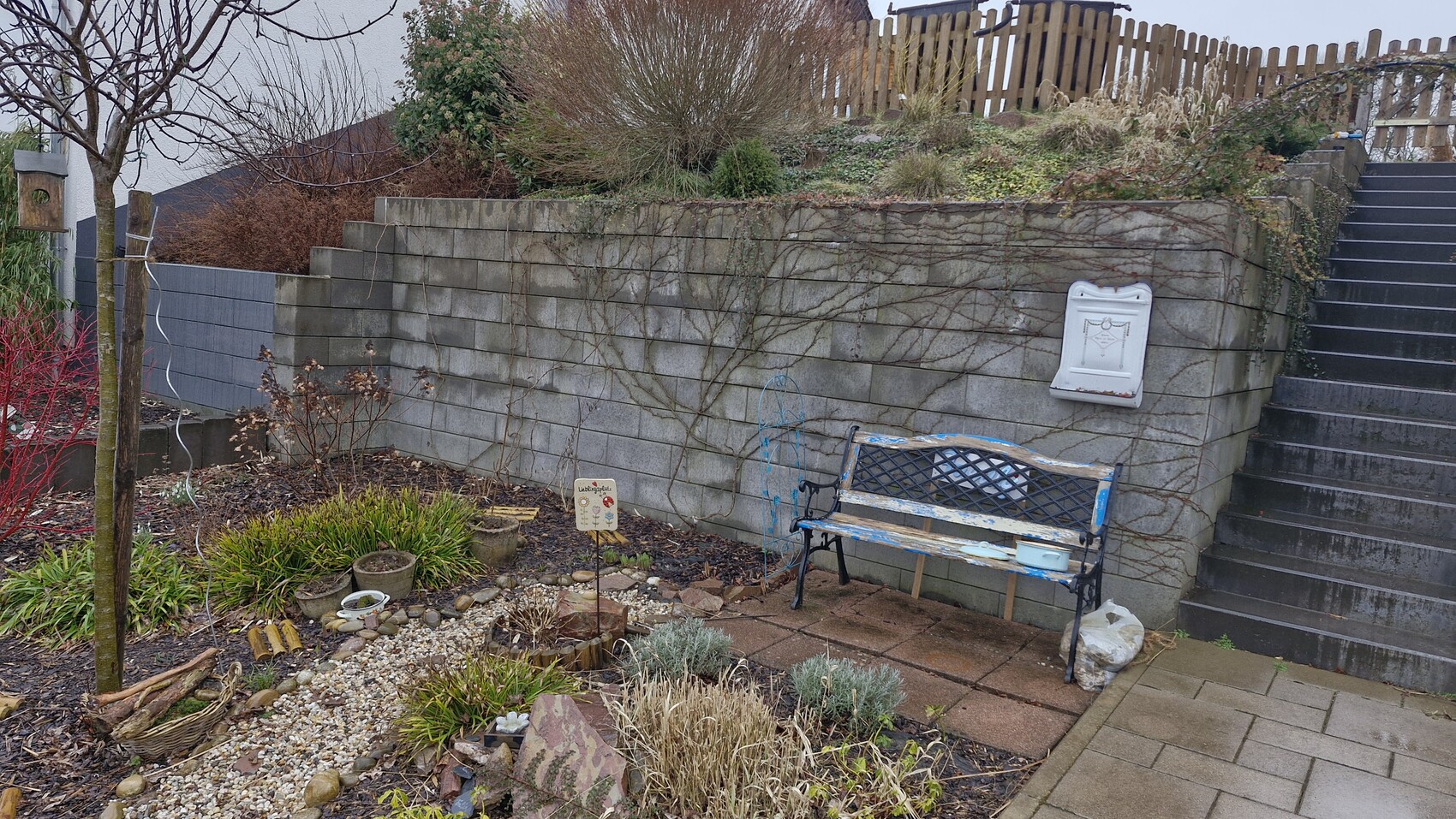Garten mit Steinmauer, Treppe rechts, alte blaue Bank, Pflanzen und weißer Briefkasten.