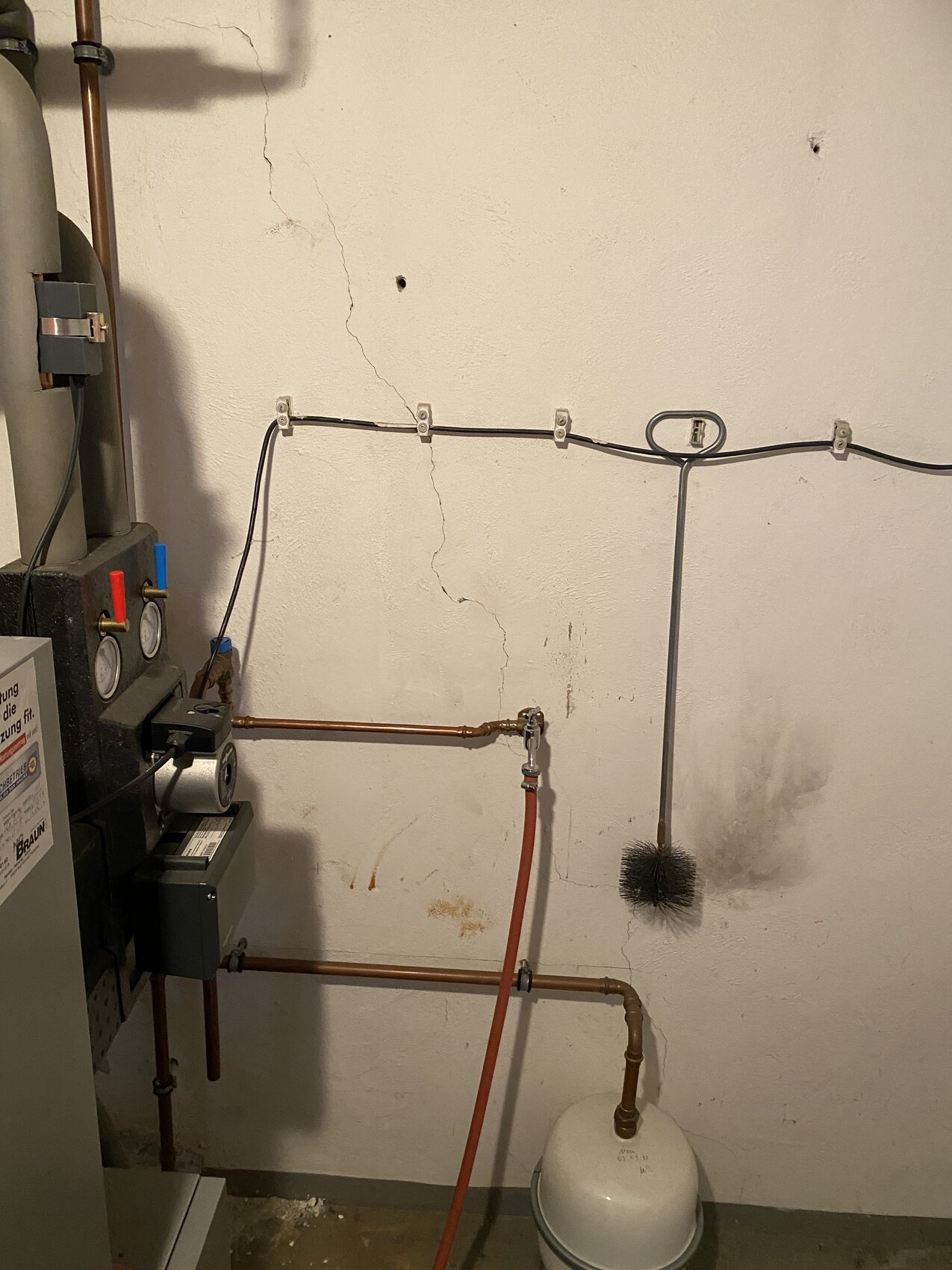 Heizungsraum mit Boiler, Kupferleitungen, rotem Schlauch und Reinigungsbürste; Wandrisse.